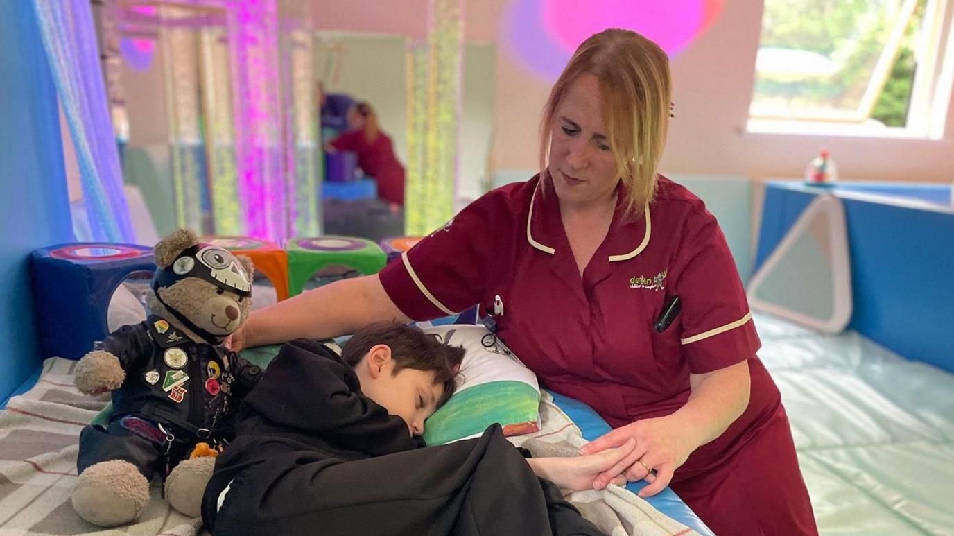 Sue holding the hand of young hospice patient as he sleeps on a bed next to a teddy bear