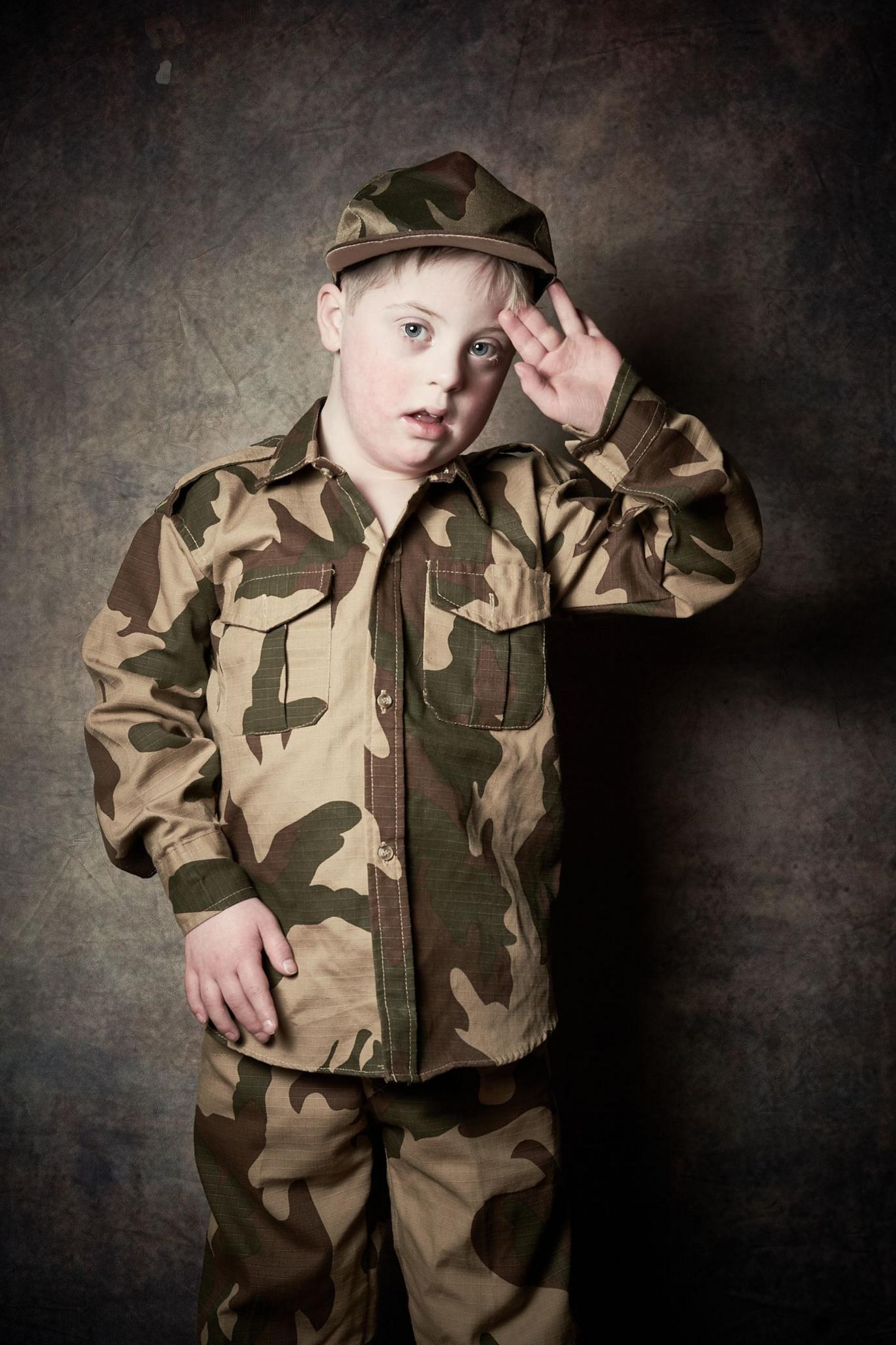 Maximus, a young boy with short blond hair salutes the camera as he wears camouflaged trousers, jacket and hat