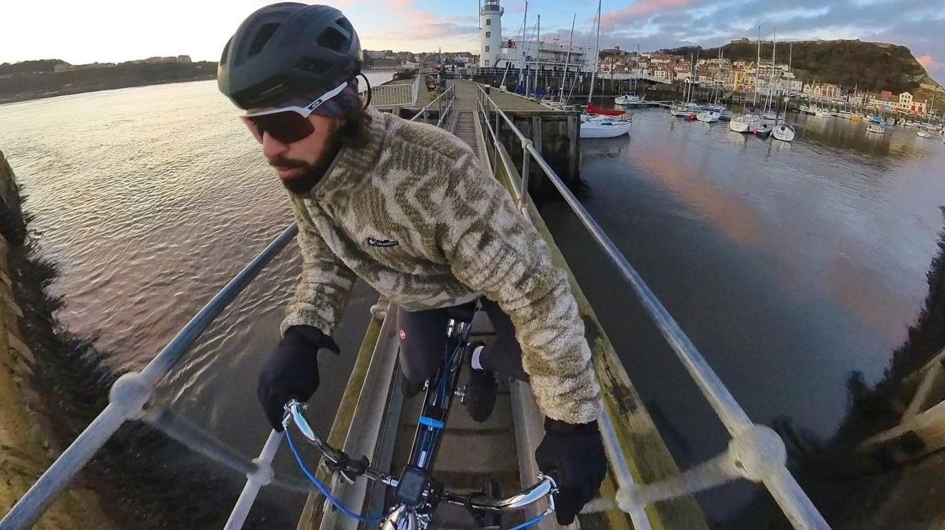 Mike Humphreys riding the Chopper along a narrow railed bridge over the water. There are boats, houses and trees in the background.