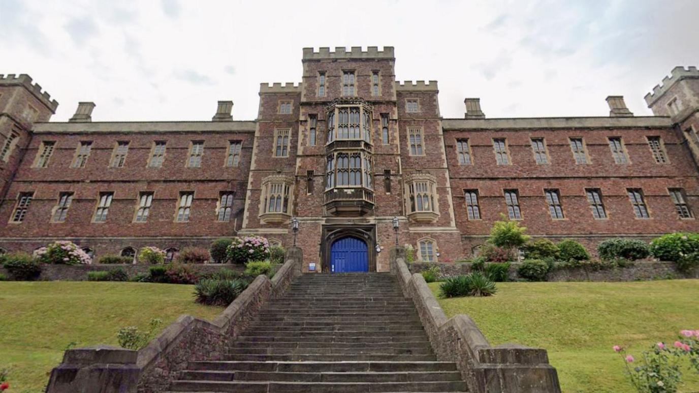 The view up to the entrance of the Queen Elizabeth’s Hospital school. It is a large 14th century brick building, with castle-like turrets, lots of windows and flowers planted outside. There is a large stone staircase leading up to a blue arched door.