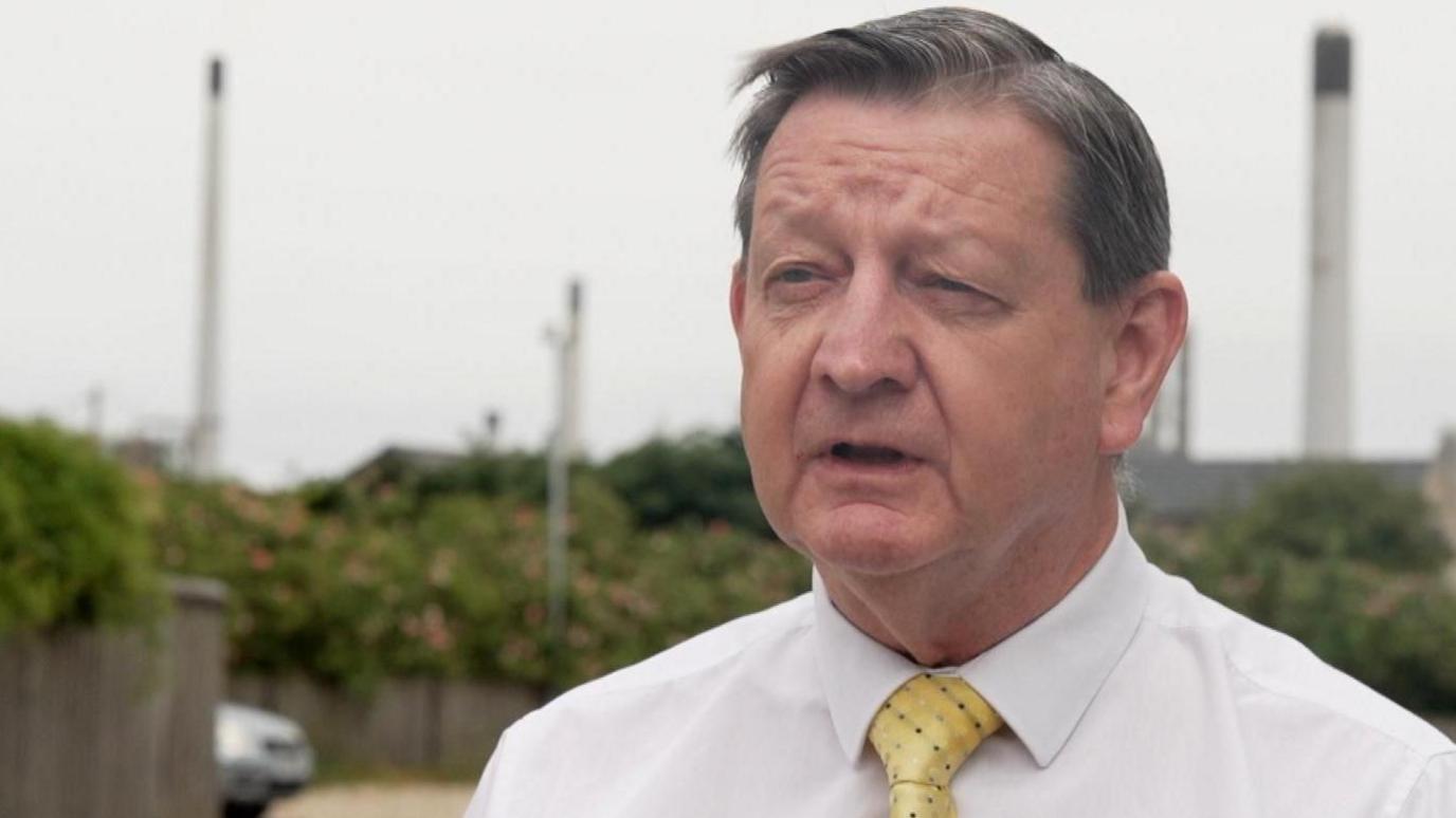 Andrew has brown eyes and short cropped dark and grey hair. He is wearing a white shirt with a yellow tie. In the background is the refinery, out of focus.