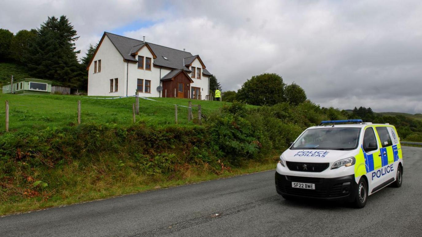 A police van is on a road below the two-storey house. There is a police officer standing in front of the property.