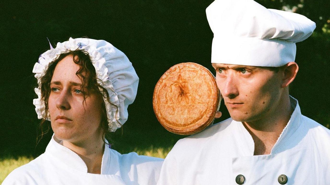 A man and a woman look into the distance wearing chef whites. A pie floats between them