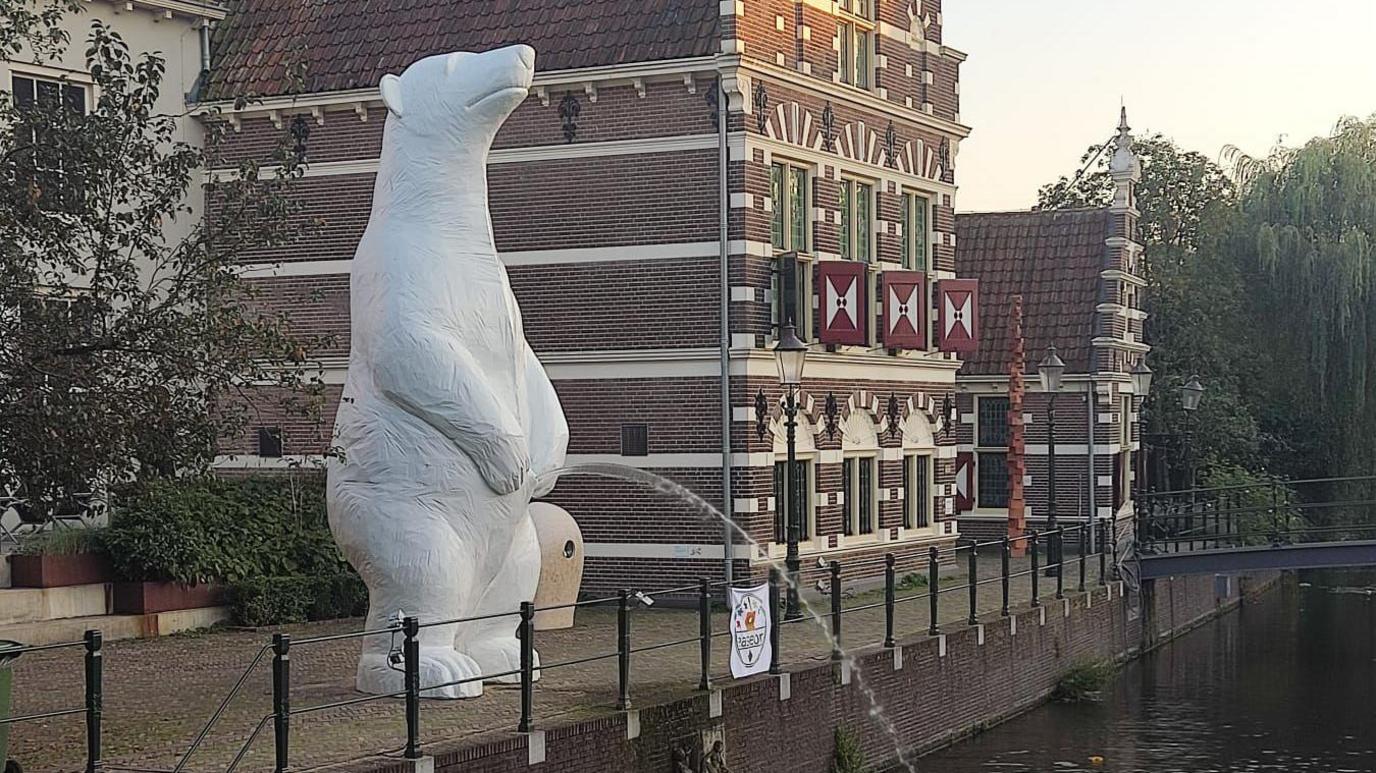 A sculpture of a 6m (nearly 20ft) tall white polar bear urinating into a canal with a stylish tall red brick building to the right.