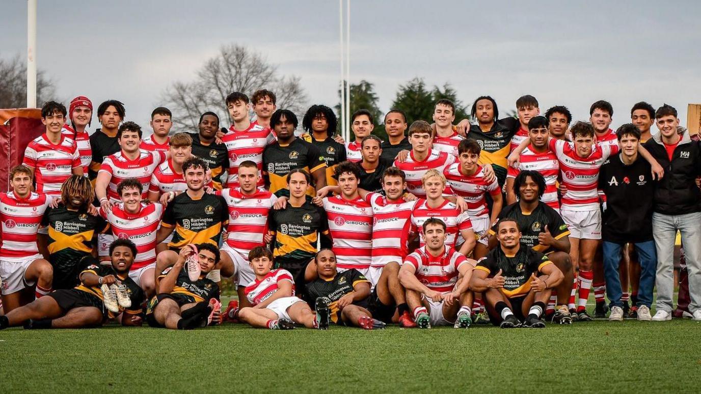 Sean Magee yn sefyll yn gwisgo cap yn y rhes gefn Mae o yn gwisgo crys rygbi a shorts tim Gibraltar. Mae aelodau'r ddau dim oedd yn chwarae'r gem yn sefyll ac yn eistedd i gael tynnu'r llun o'r timau ar ddiwedd y gem.