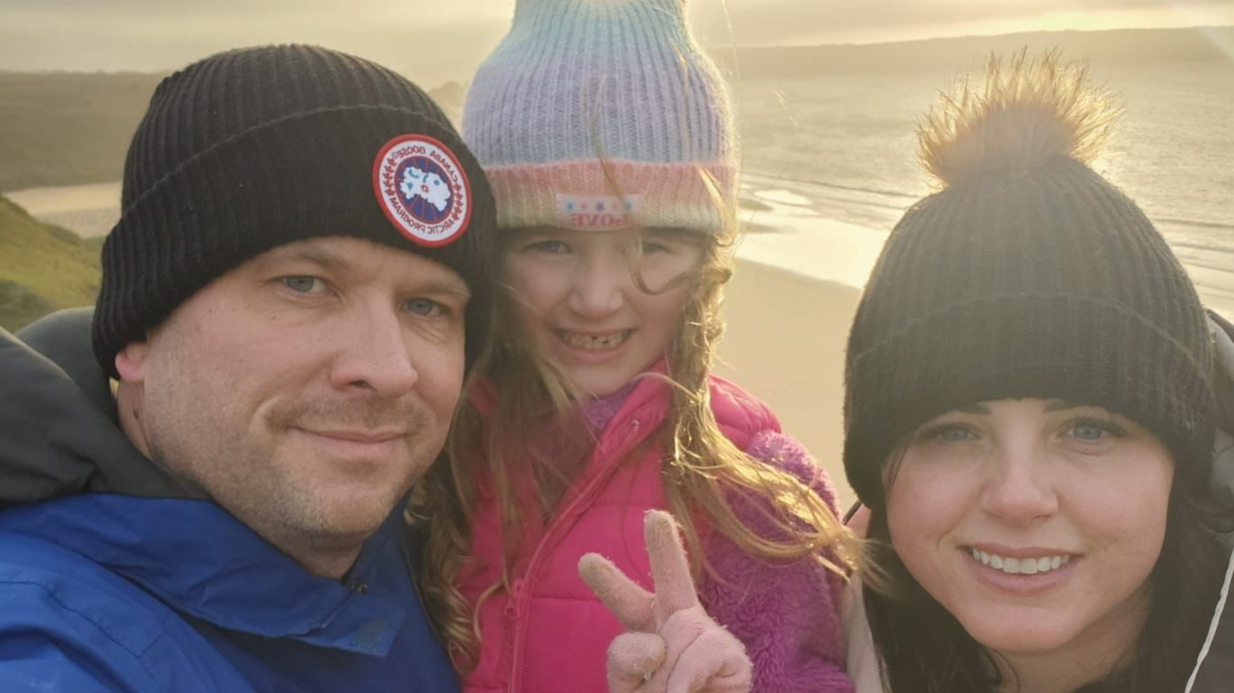 Angharad Dennis smiles as she takes a selfie with her husband and daughter on a beach. They wear hats and coats with the sun behind them.