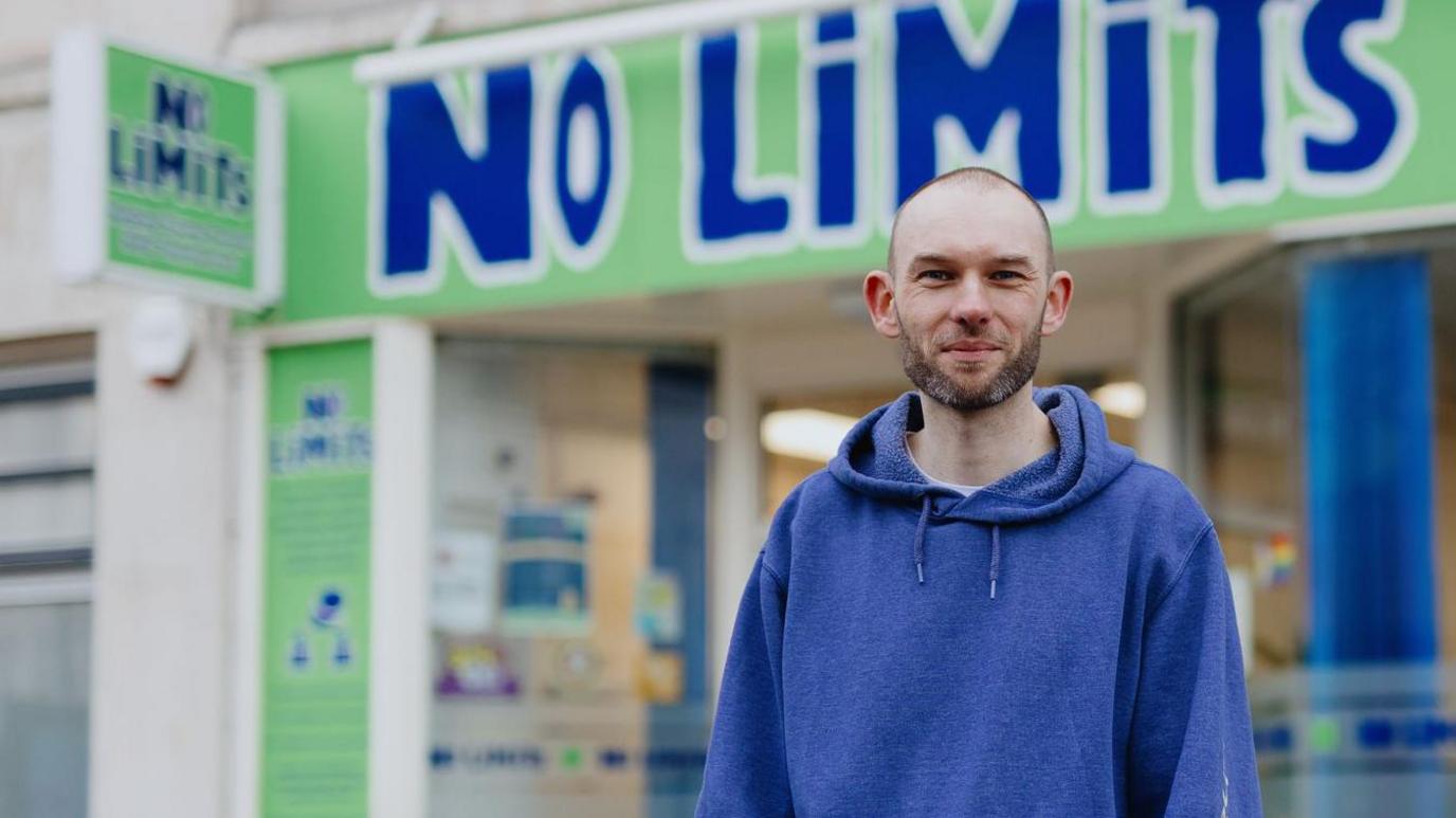 James is bald with a brown and grey stubble. He is wearing a blue hoodie. Out of focus behind him is a building with large green and blue signage saying 'No Limits'.