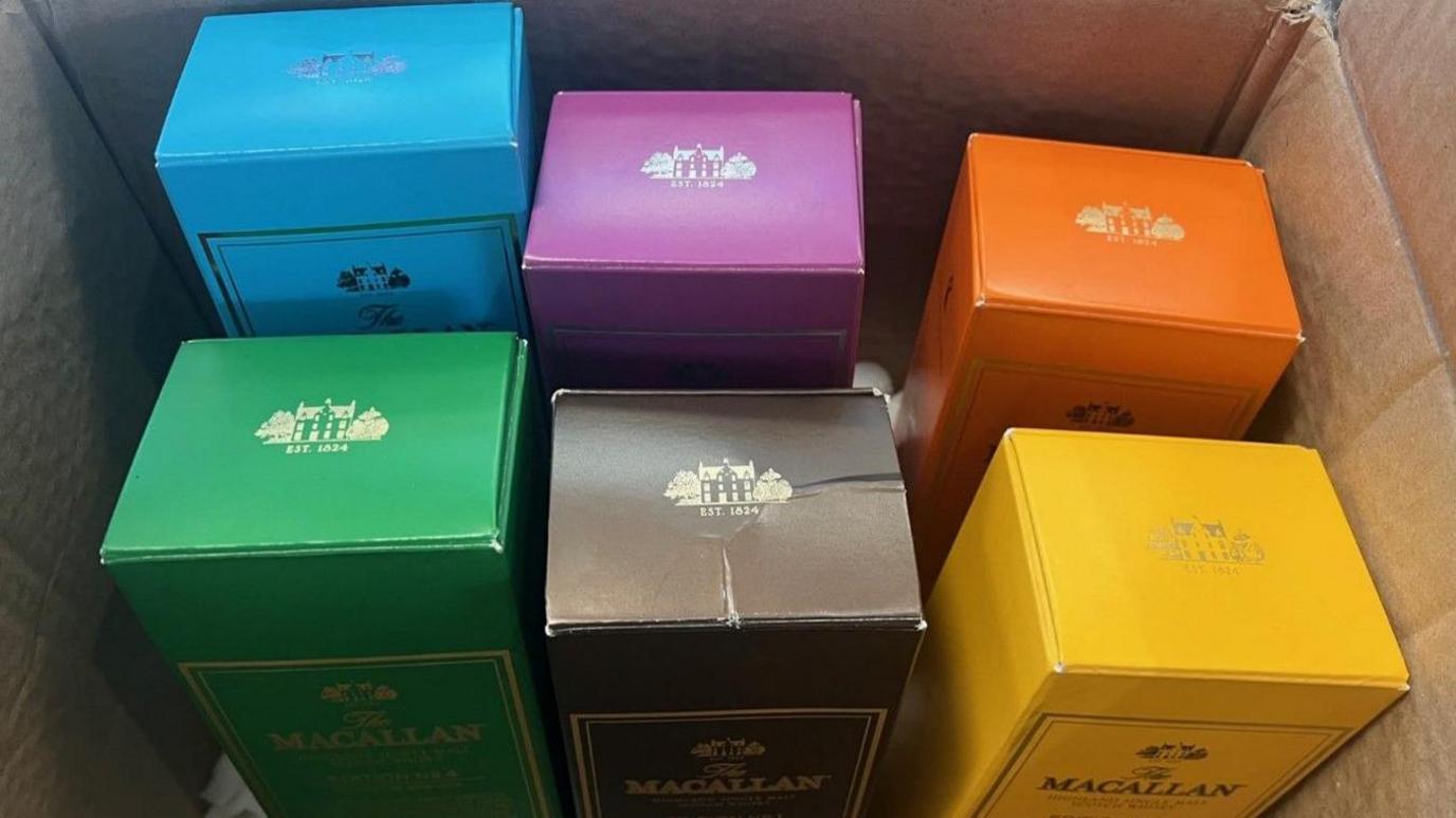 Six brightly-coloured boxes for Macallan whisky, one in each of the colours blue, yellow, orange, pink, green and brown, stand in a cardboard box