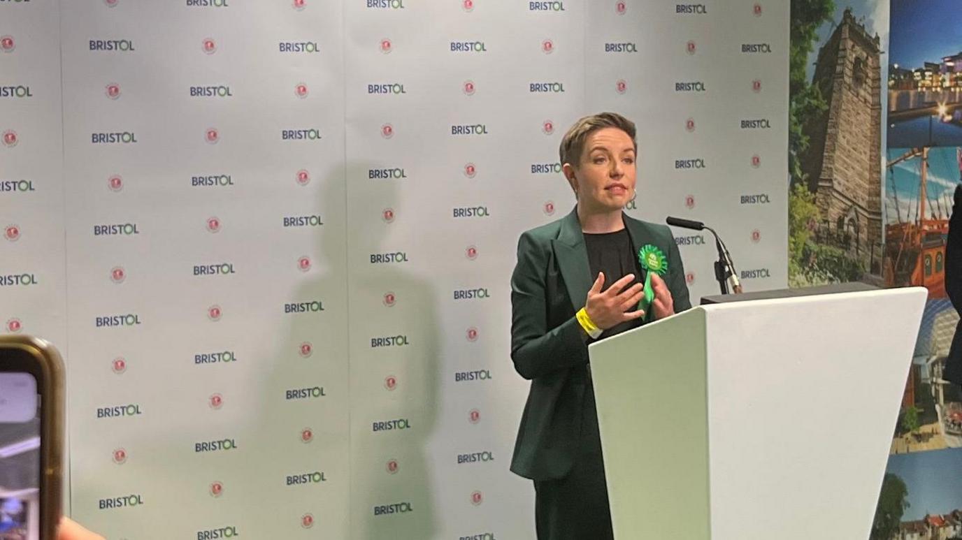 Carla Denyer standing in front of a podium wearing a green suit and rosette