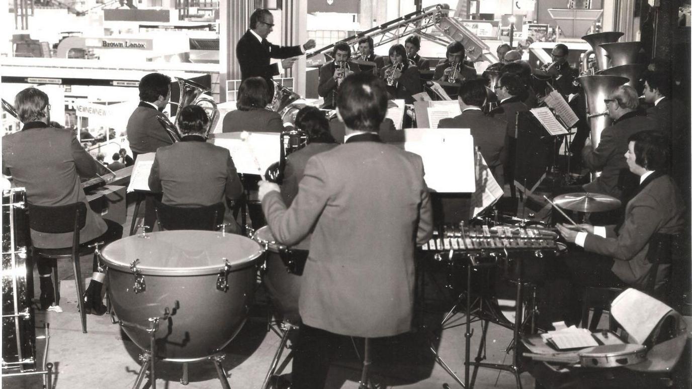 A black and white picture of a brass band performing. Musicians can be seen playing instruments as a conductor points at them.