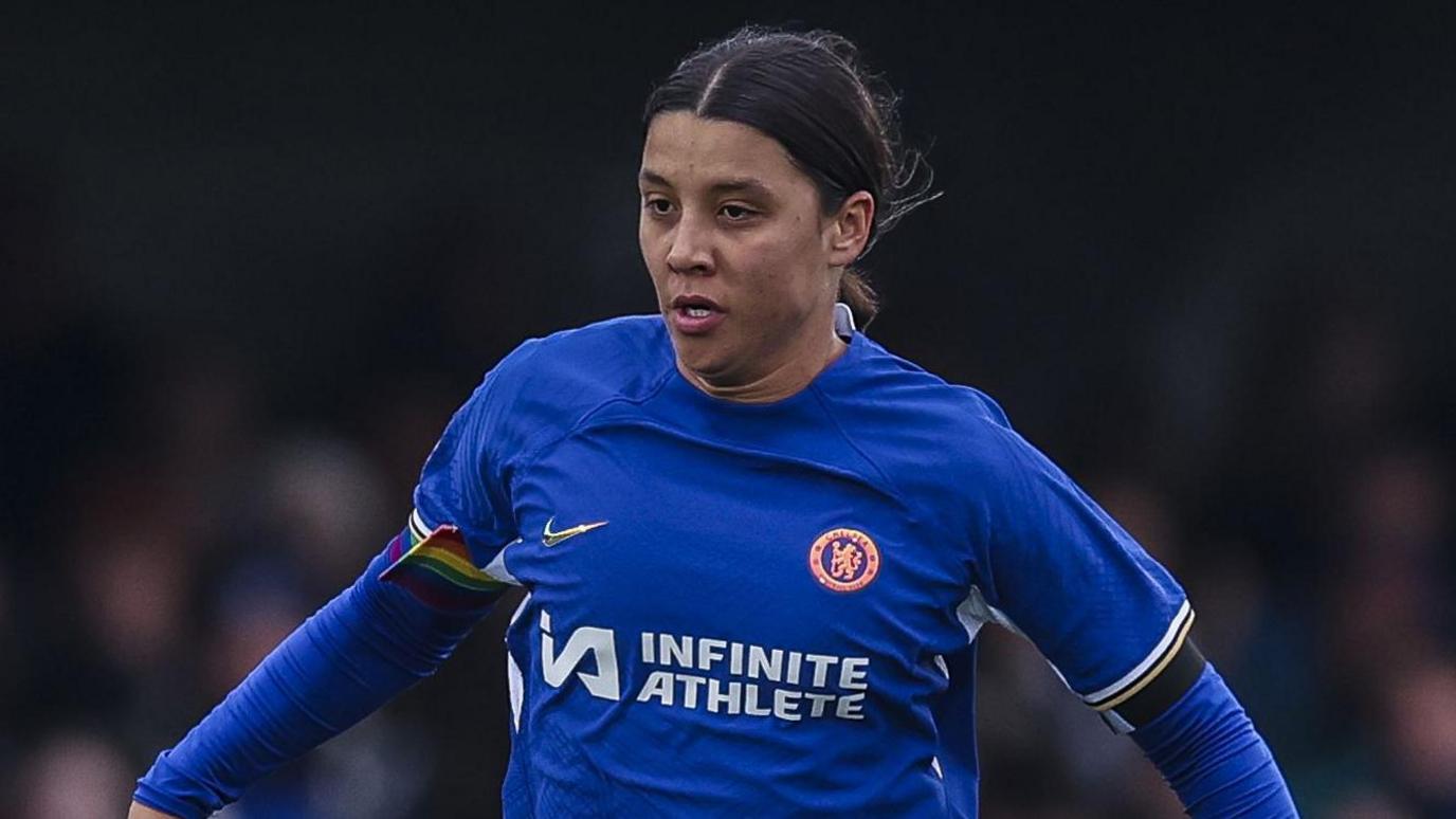 Sam Kerr on the pitch playing football. She wears a blue Chelsea football top with the branding 'Infinite Athelete' and her brown hair is tied back as she runs forwards.