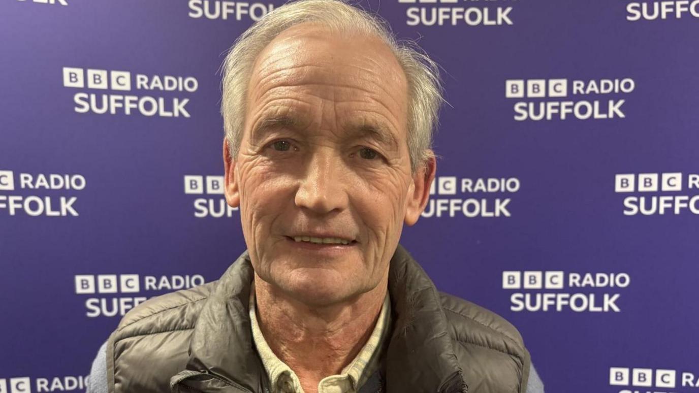 George Burley wearing a black puffer gillet with a zipped blue jumper and checked shirt underneath, standing in front of a BBC Radio Suffolk background.