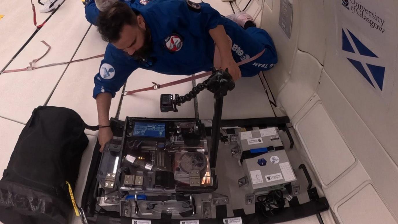 Dr Gilles Bailet, wearing a blue flight suit, tests a 3D printer while strapped into a zero gravity aircraft. There is a saltire flag and the University of Glasgow logo taped to the wall of the aircraft.