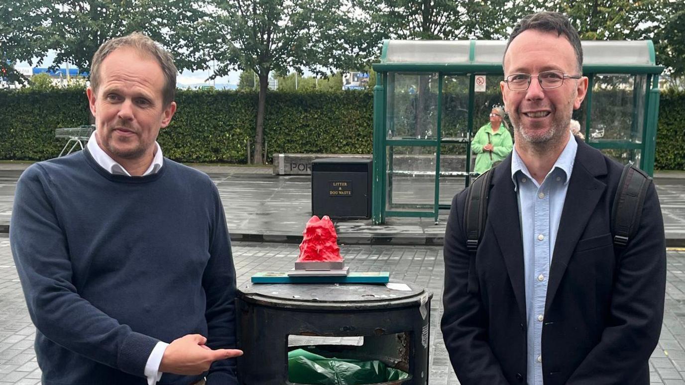 Two men stand by a bin with a DIY award on top of the bin. The award is a red textured object on a wooden plank. One man points at the opening to the bin.