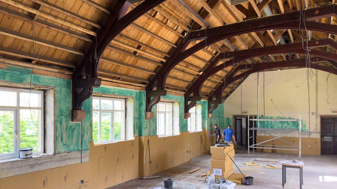 Workmen inside the hall during the recent refurbishment work. There is scaffolding up, the ceiling has been pulled back to its bare beams, and the new ecological green-coloured insulation is visible on the walls around the large windows.