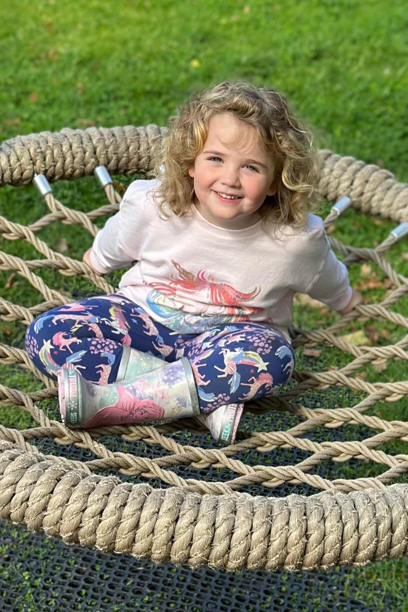 Amber is sitting on a wide circular rope seat which has rubber matting below it and grass beyond it. She is smiling at the camera. Her arms are outstretched behind her and her legs are crossed in front of her. She has shoulder-length blonde curly hair and clue eyes. She is wearing a brightly coloured sweatshirt with a unicorn on it, unicorn-printed leggings and unicorn-printed wellies.