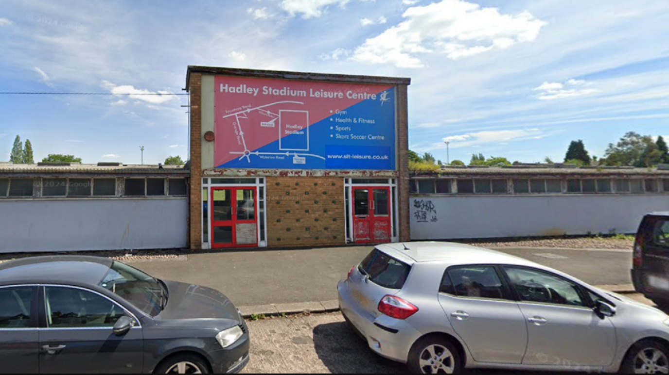 A low building, which looks slightly run-down, next to a road where cars are parked. In the middle of the building is an entrance above which is a sign reading "Hadley Stadium Leisure Centre".