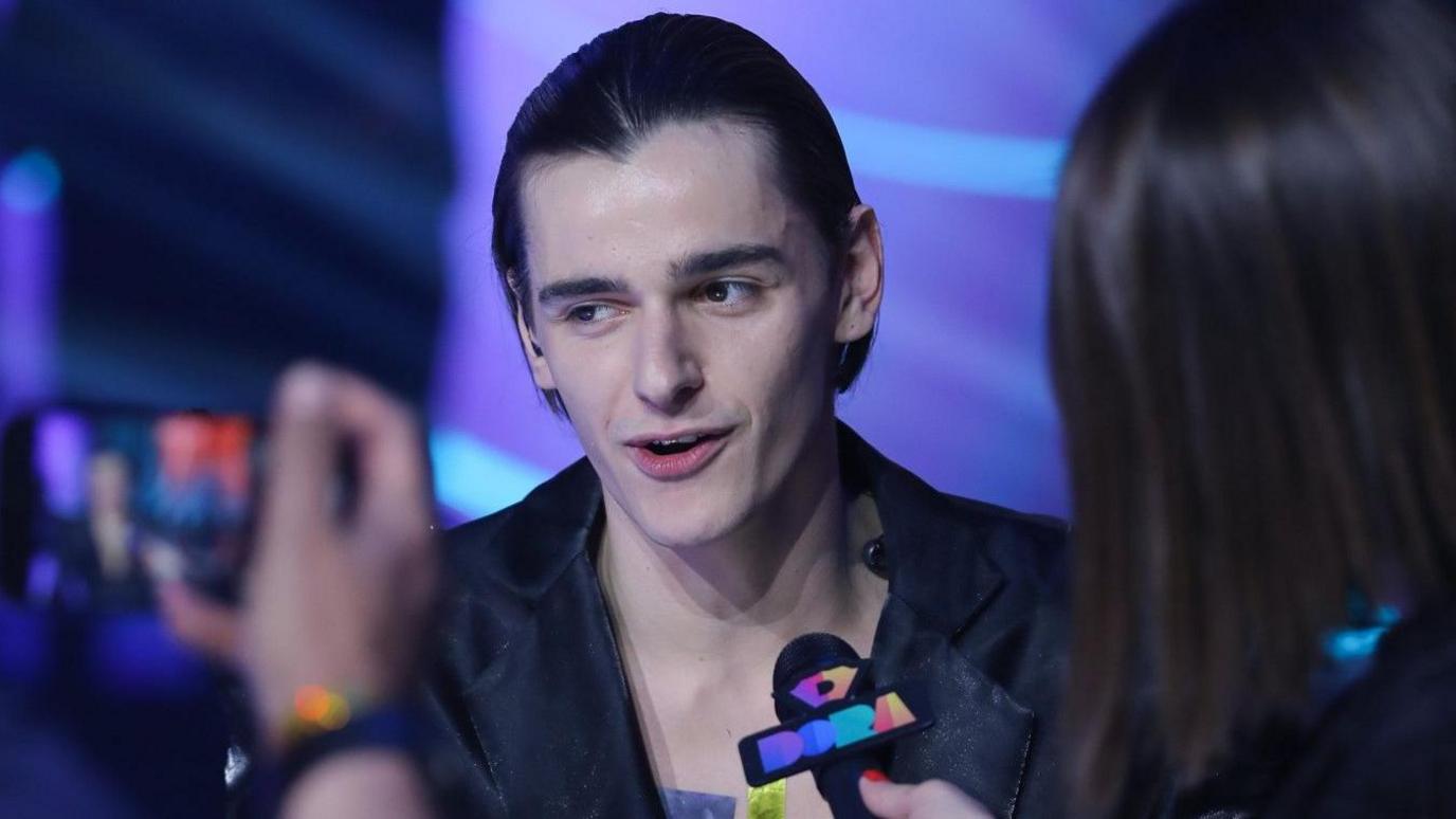 Marko Bosnjiak surrounded by reporters and photographers at the end of the Dora contest. He is dressed in a black jacket and has slicked back black hair.