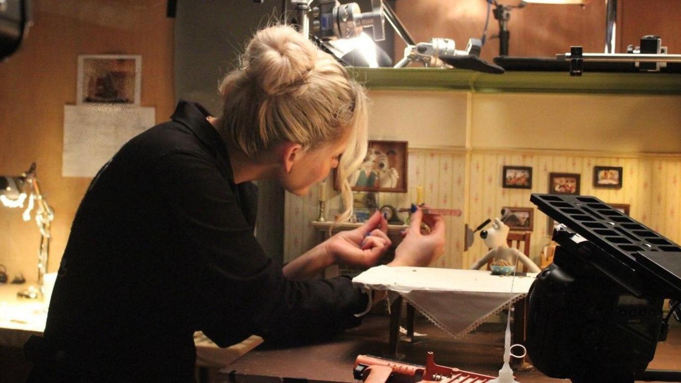 Beth Jones crouches over a miniature set of a sitting room and places a small prop on the mantlepiece. Beth is wearing black and has blonde hair which is tied back in a bun. There are lights pointed towards the set as she works. 