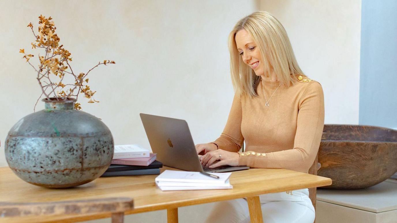 A woman at her desk. She has blonde hair and is typing on a laptop. She is wearing a camel-coloured t-shirt. Next to the laptop is a large grey vase with light brown foliage in it. 