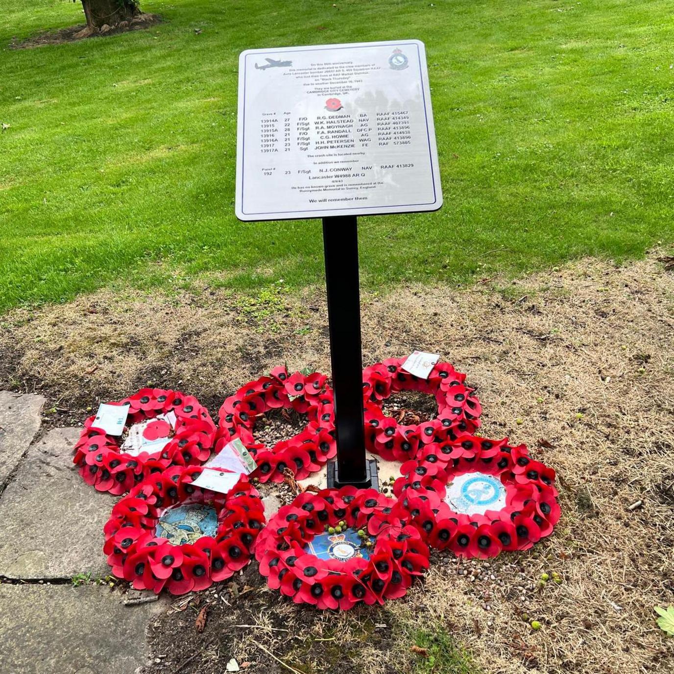 Six poppy-wreaths surround the base of a plaque on a patch of land in a village. The rectangular, light-grey plaque lists information and names and is mounted on a black stand.