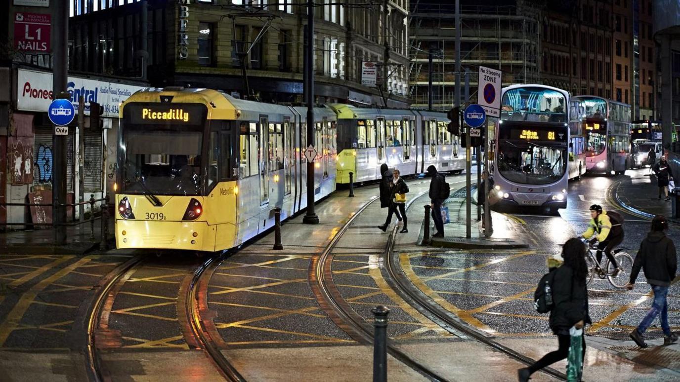 People walk by a tram and a bus at Shudehill stop in the evening at Manchester city centre