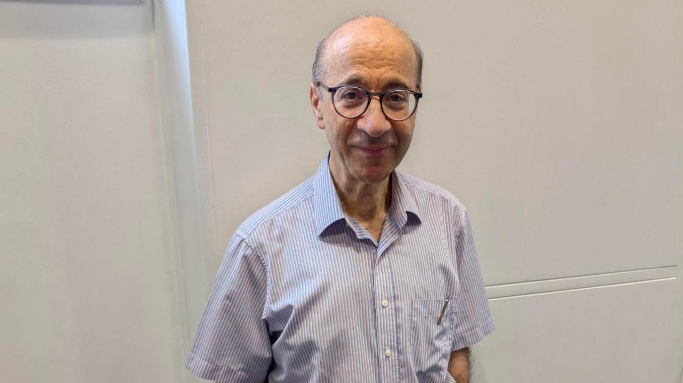 Paul Harris standing by a white wall, with short hair, glasses and wearing a stripped shirt.