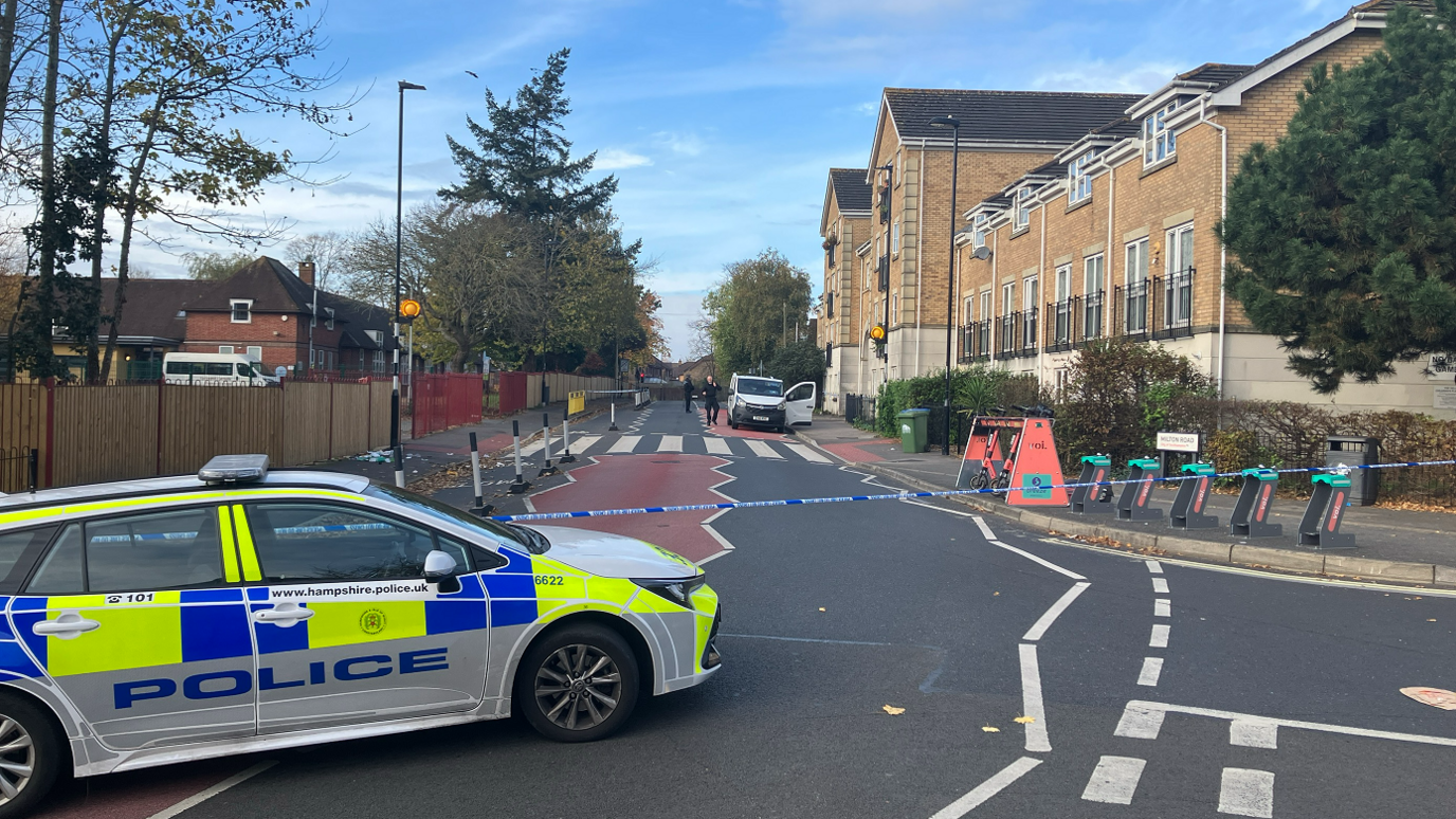 A police car is parked on Milton Road, with police tape cordoning it off to people. There is a white van parked further up the road with people who seem to be police officers walking around it 