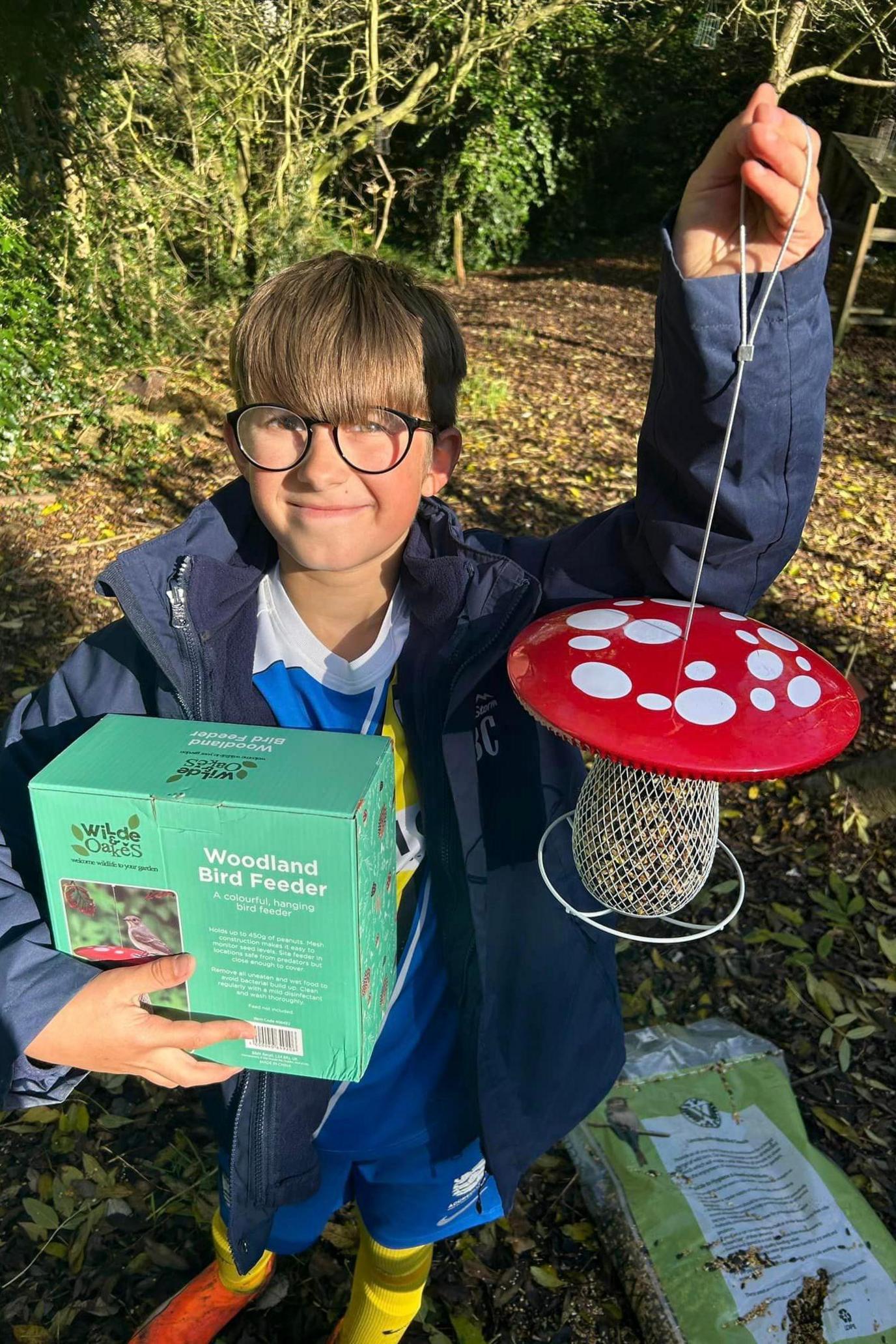 A boy with a brown fringe and large glasses holds up a bird feeder shaped like a red toadstool. 