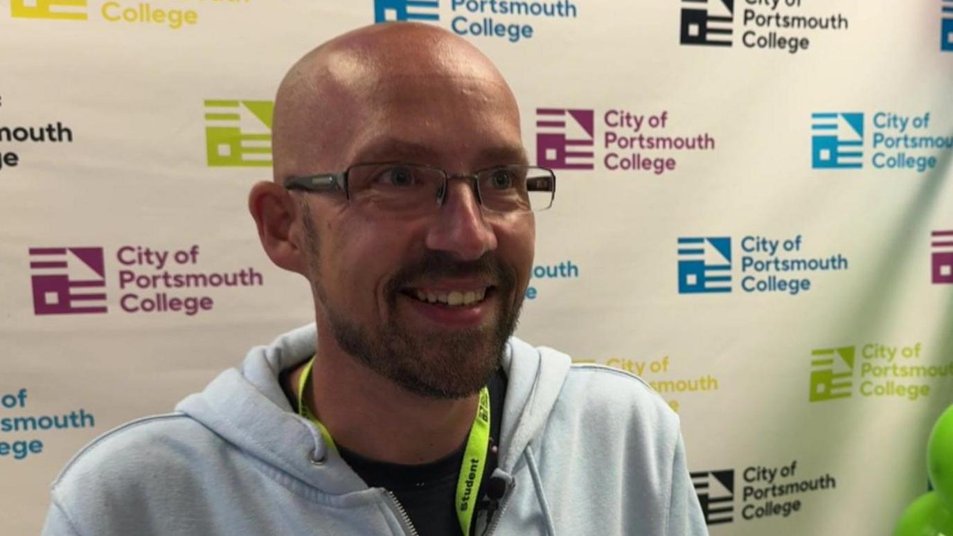 A bald man wearing glasses is grinning. He is standing in front of a screen which has the City of College Portsmouth logo on.