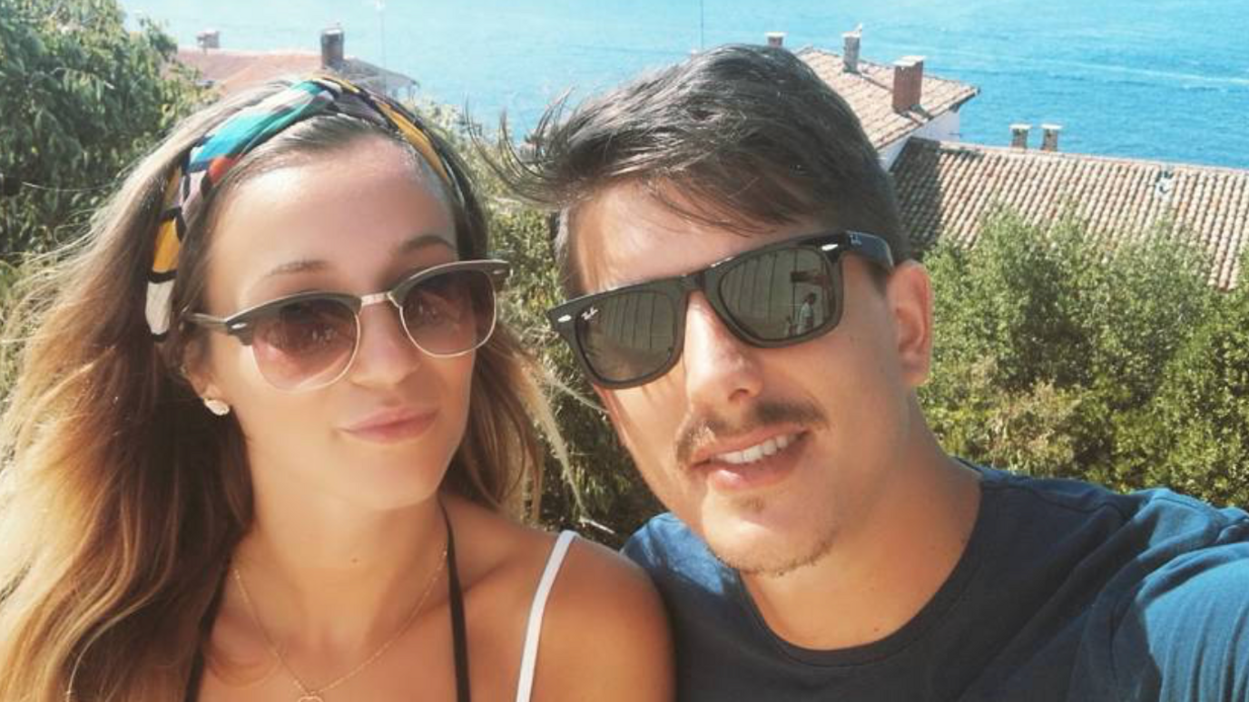 Young couple, one woman with medium brown hair and a young man, with short dark brown hair and a moustache both wearing sunglasses and looking at the camera taking a selfie. She has thin straps for a top and he is wearing a blue t-shirt - behind them are rooftops and the sea. 