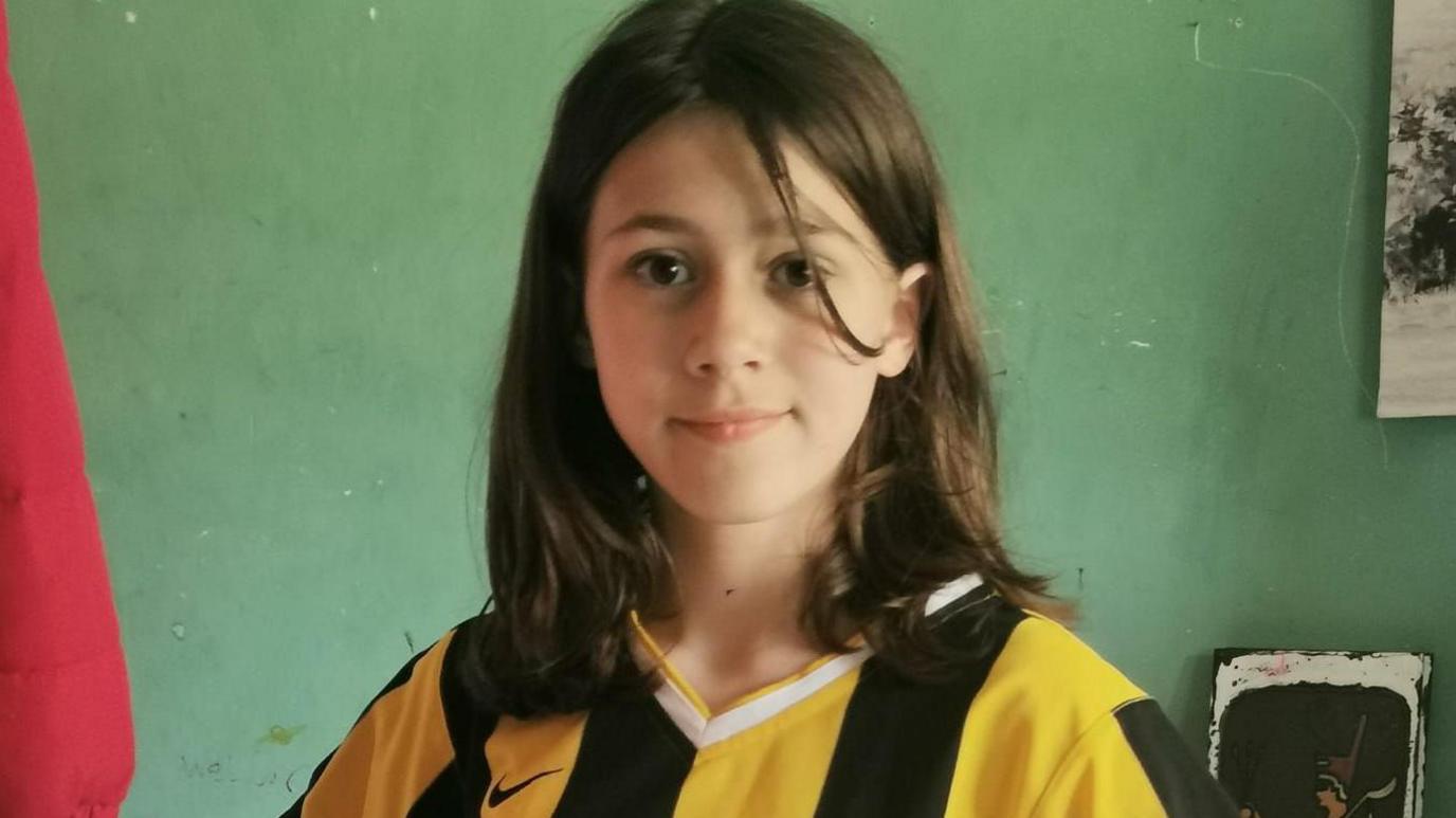 A young girl with brown hair, smiling for the camera while wearing a striped yellow and black football top