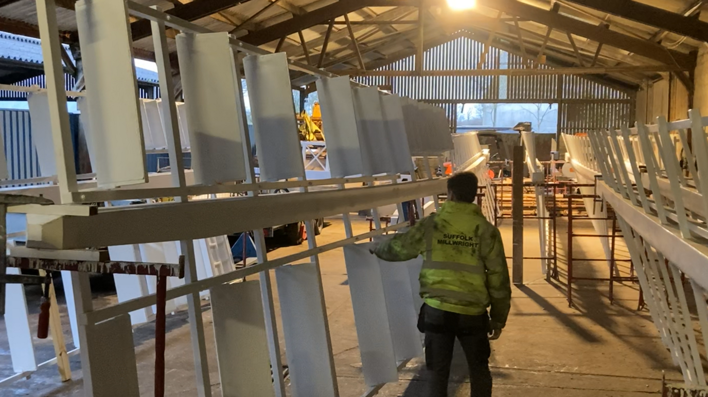 Mr Whiting walks through his warehouse and reaches out to a large white windmill sail. He has his back to the camera and he wears a florescent green hi-vis coat.