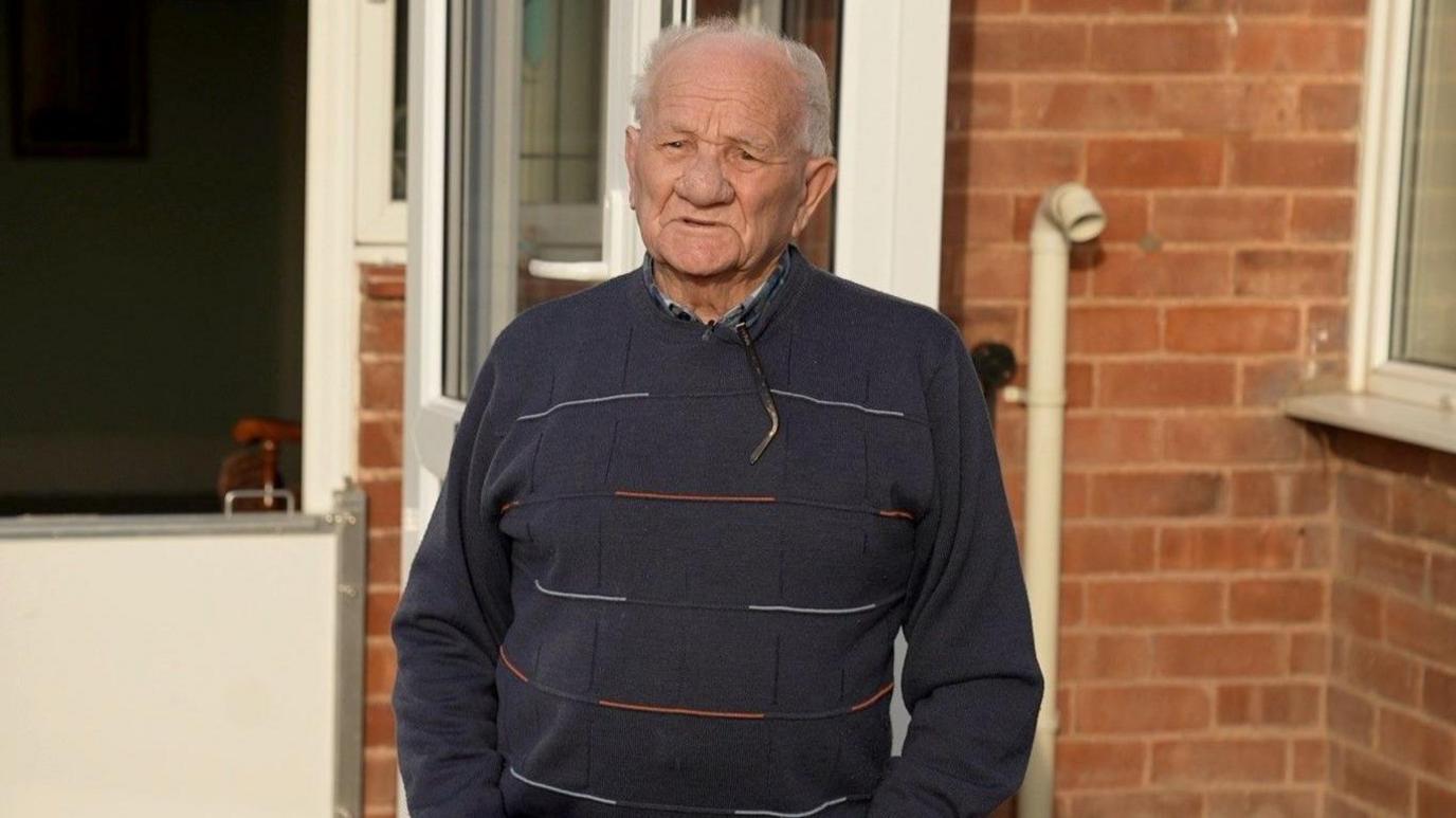 An older man, who has very short grey hair standing outside of a house. He is wearing a navy jumper with white and red embroidery. 