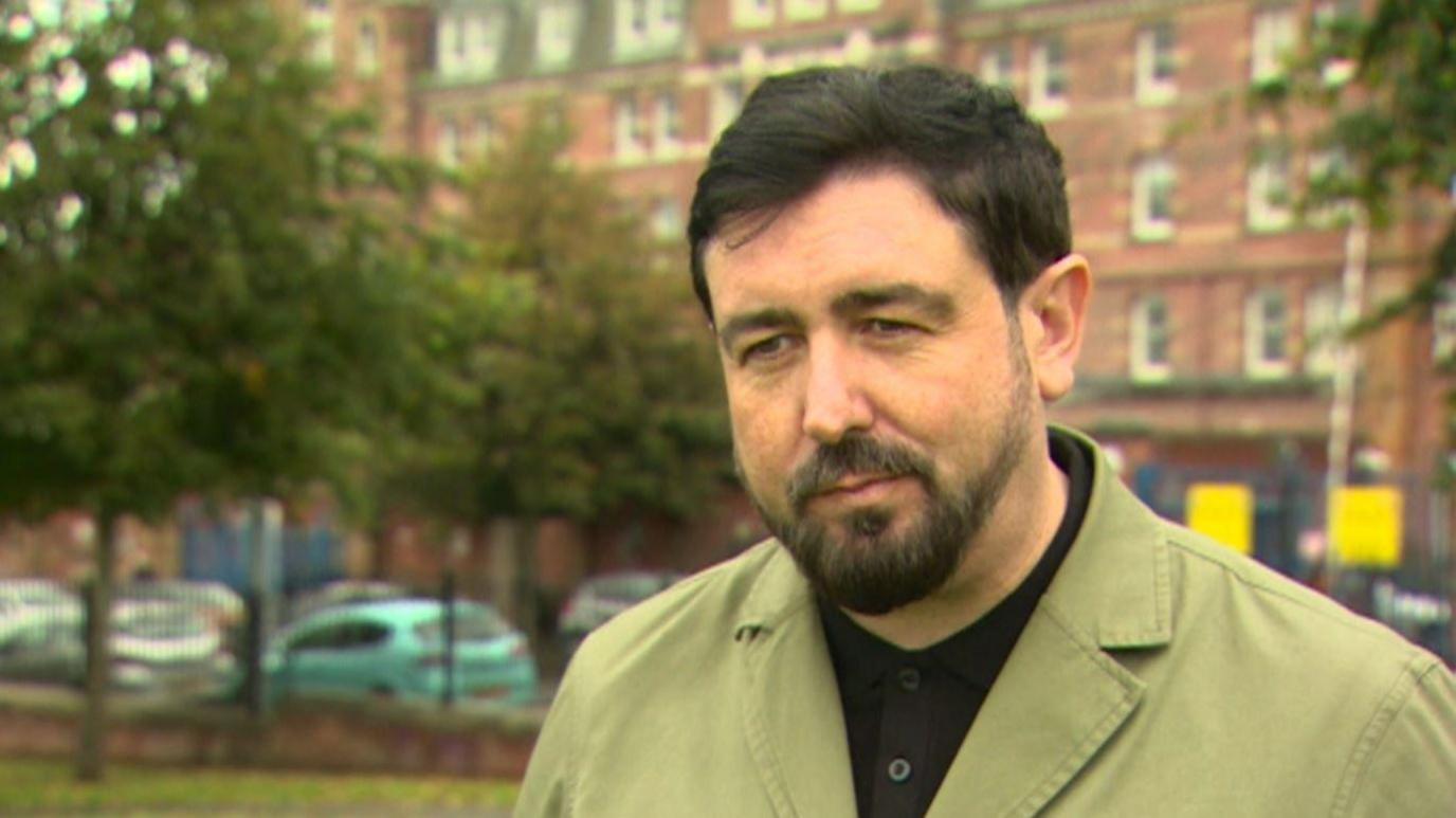 Paul Doherty standing in front of the Royal wearing a black shirt and green coat, with black hair and beard