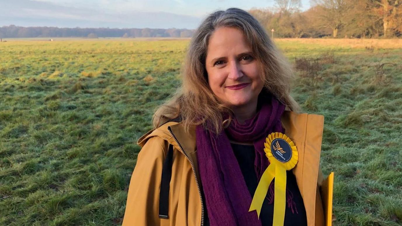 Lara Pringle smiles at the camera. She has dark brown hair and is wearing a yellow jacket and a purple scarf, with a Lib Dem rosette on it. She is standing in a field with dense woodland in the background.