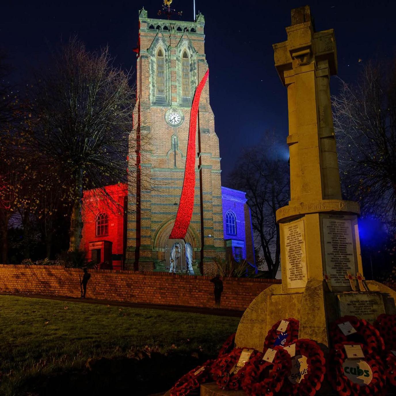 A church with a cascade of knitted poppies on its front, seen from a distance appearing as a streak of red over the church. There is a war memorial in the forefront of the picture with wreaths of poppies at its base.