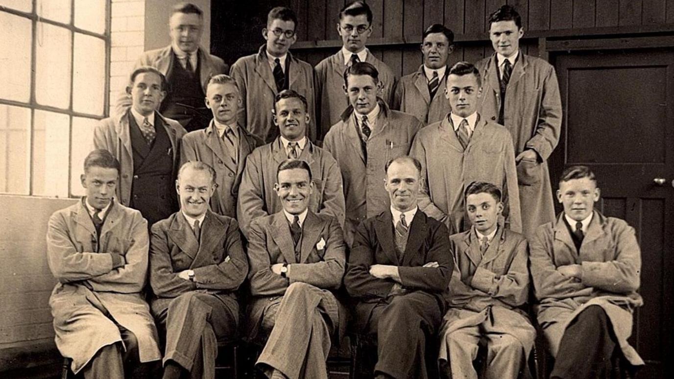 A group of Post Office engineers from the 1940s, in black and white. They are all wearing long overcoats, ties and white shirts. Fred Martin is on the back row wearing glasses and staring at the camera. Many of the men are also smiling.