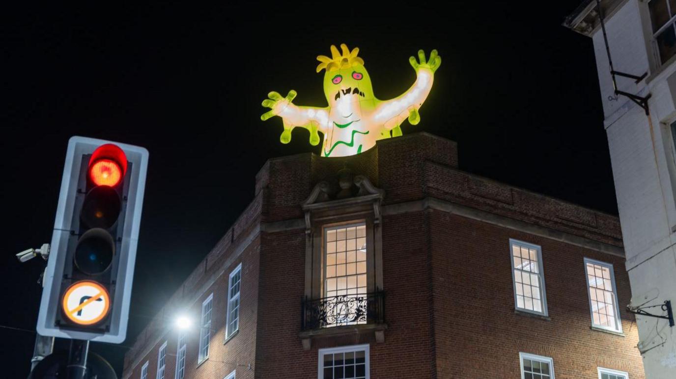 A slimy green enormous inflatable monster with its arms up above a building. Traffic lights are in the bottom left corner of the photo.