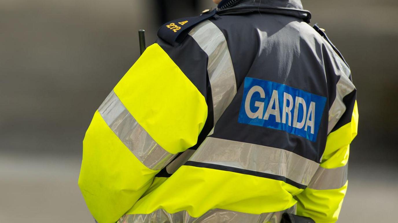 The torso of a female garda officer on duty on a street. She is wearing a yellow and navy waterproof high viz  jacket with silver reflective stripes, navy and gold epaulettes and the word "Garda" in capital letters on her back.