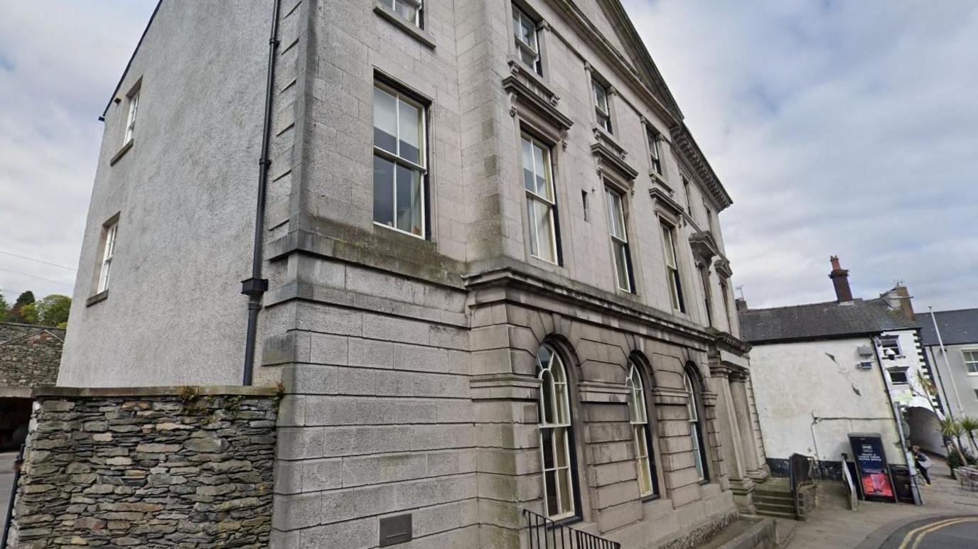 The Bank Building in Ulverston. It is a large grey stone building, three storeys high with a pointed front reminiscent of Roman or Greek architecture. There are three arched sash windows on the ground floor, with five rectangular ones on each of the upper floors. The gable end is mainly rendered in grey, but there are two smaller windows towards the rear, on on each of the upper floors. Stone steps with a black railing lead up to the entrance at the far end.