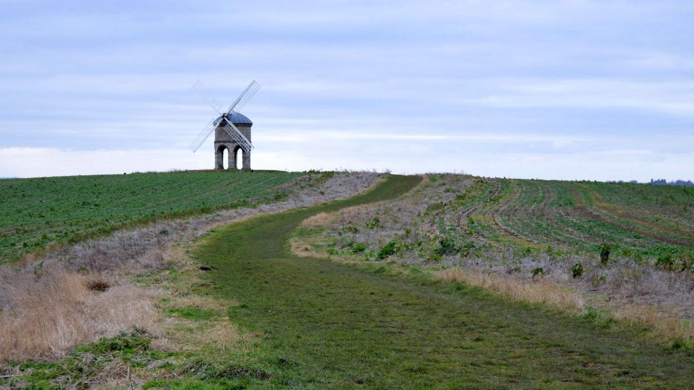 A photograph of Chesterton Windmill with four white sails. In the foreground are green fields.