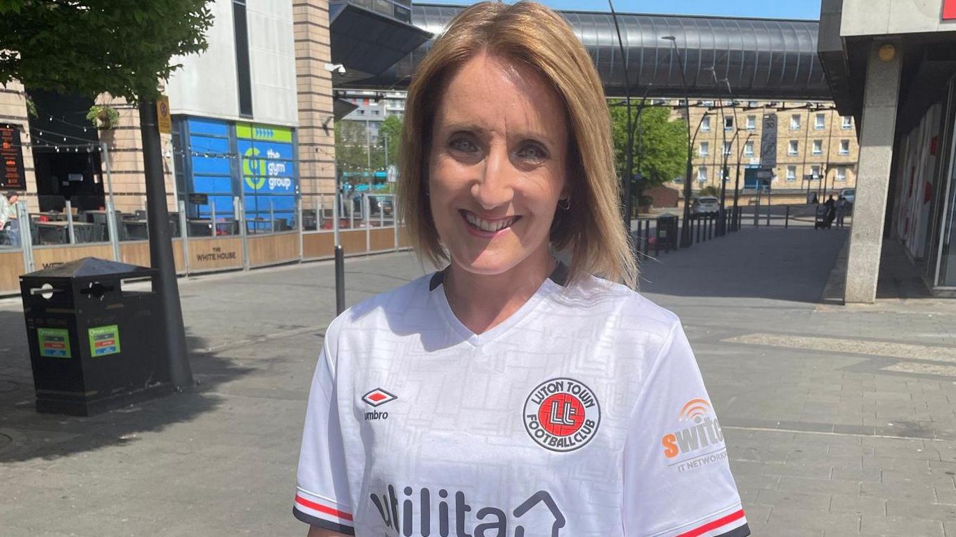 Helen smiles at the camera standing in St George's Square outside the library. There are buildings and a tree behind her. She wears a white v-neck Luton Town shirt with Utilita, Umbro and Switch advertising on.