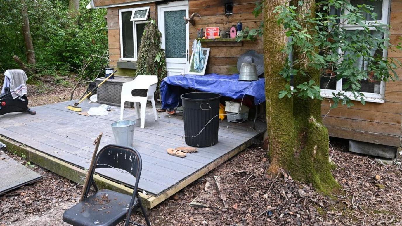 There is a wooden cabin with windows and wooden decking in front of it.
There is a black chair in the foreground, with a white chair on the decks and a table covered by blue plastic, beside a mature tree.