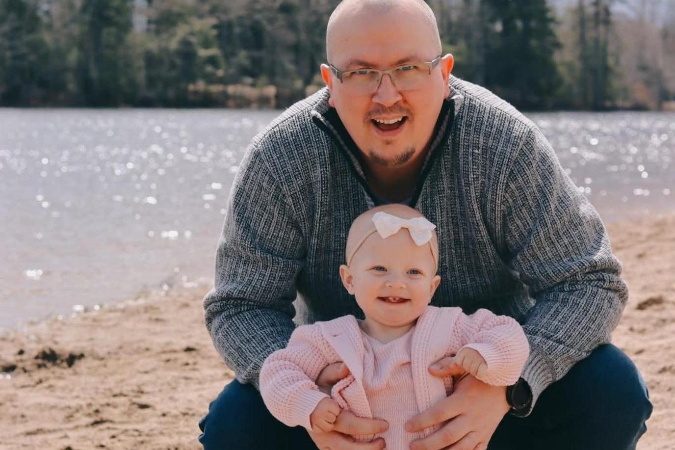 Chris Wade with a bald head and glasses wearing a light blue striped jumper. He is crouched down on a beach with a body of water behind him shimmering in the sun. In his arms is Layla, who is smiling. She is wearing a pink outfit and has a white bow on her head.