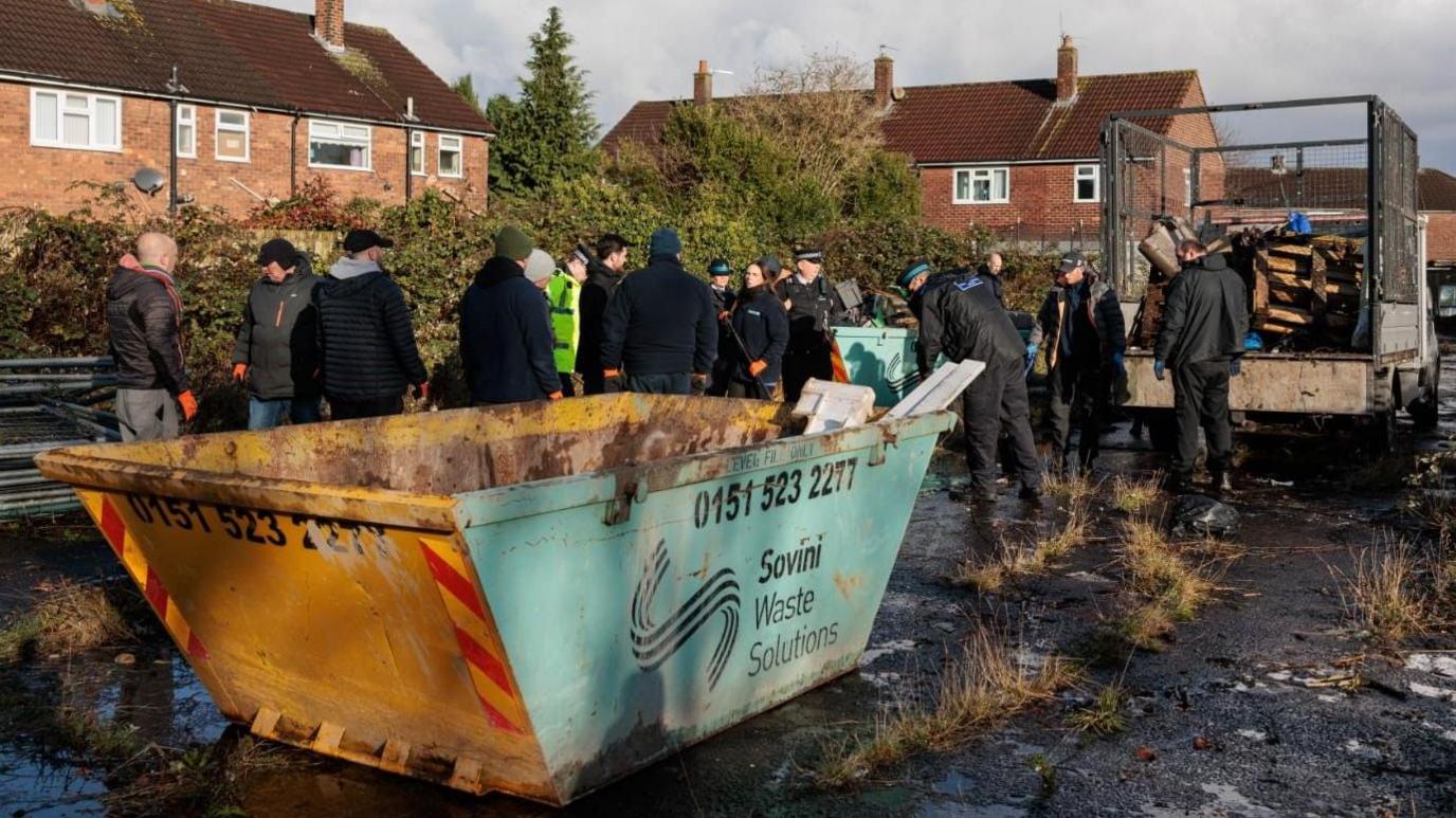 A group of approximately 20 people are stood around a blue skip