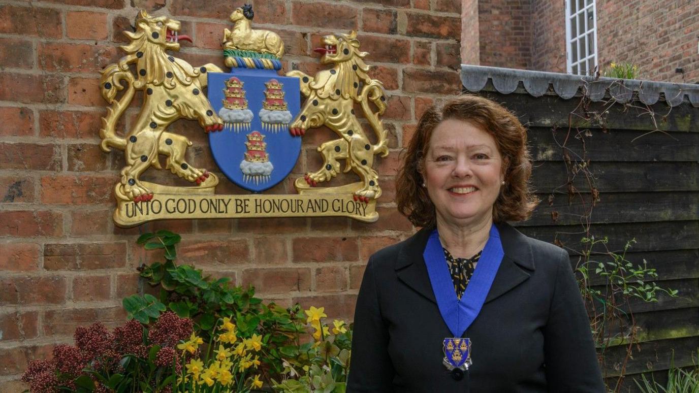 Gaynor Bowen with short brown hair smiling and wearing a black suit and a blue and gold medal on a thick blue ribbon standing outside Drapers' Hall with a gold and blue crest on the wall behind her
