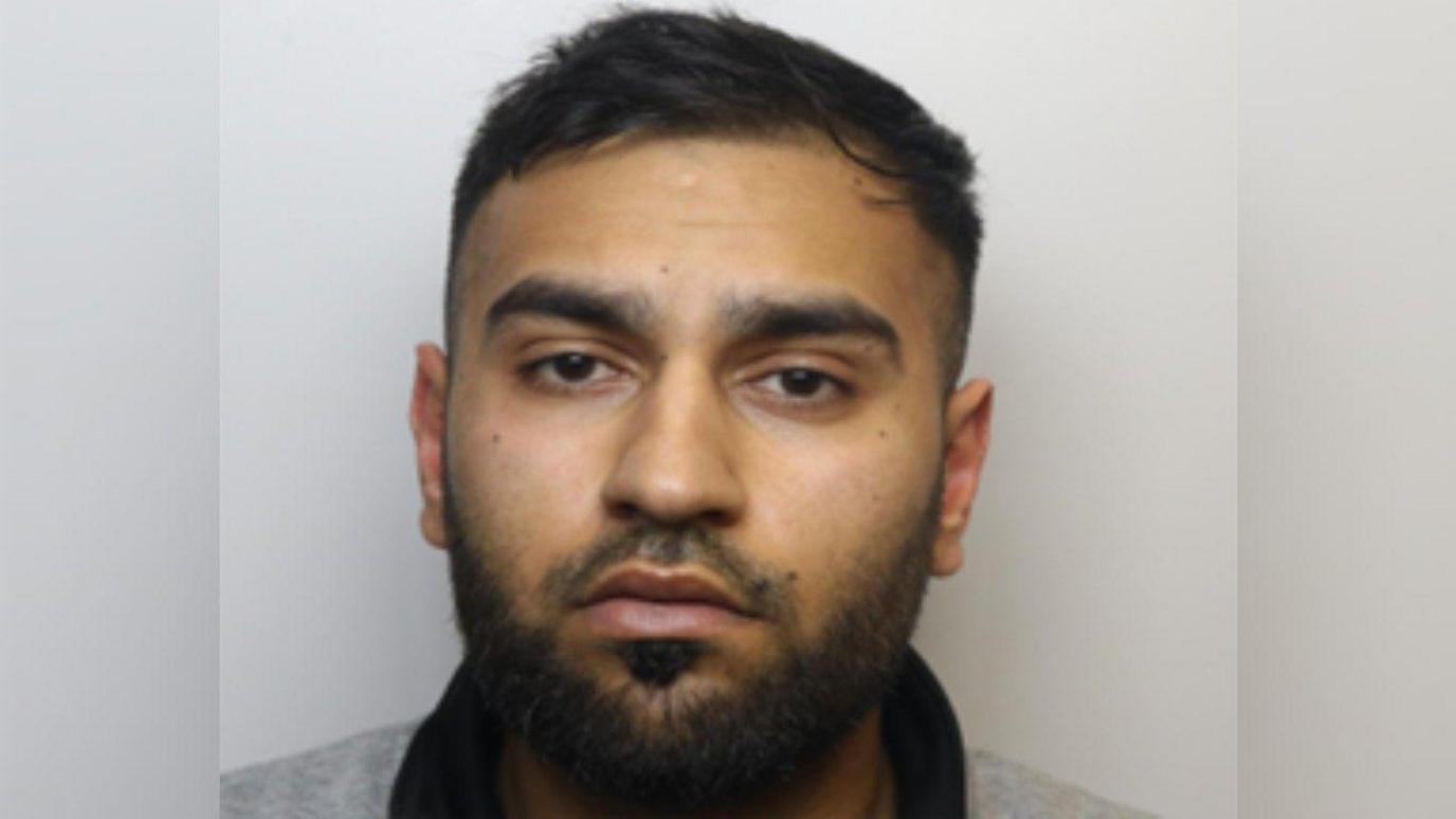 A police custody image of Zaheer Ahmed. He has short black hair and a short black beard and is wearing a grey jumper with a black collar. 