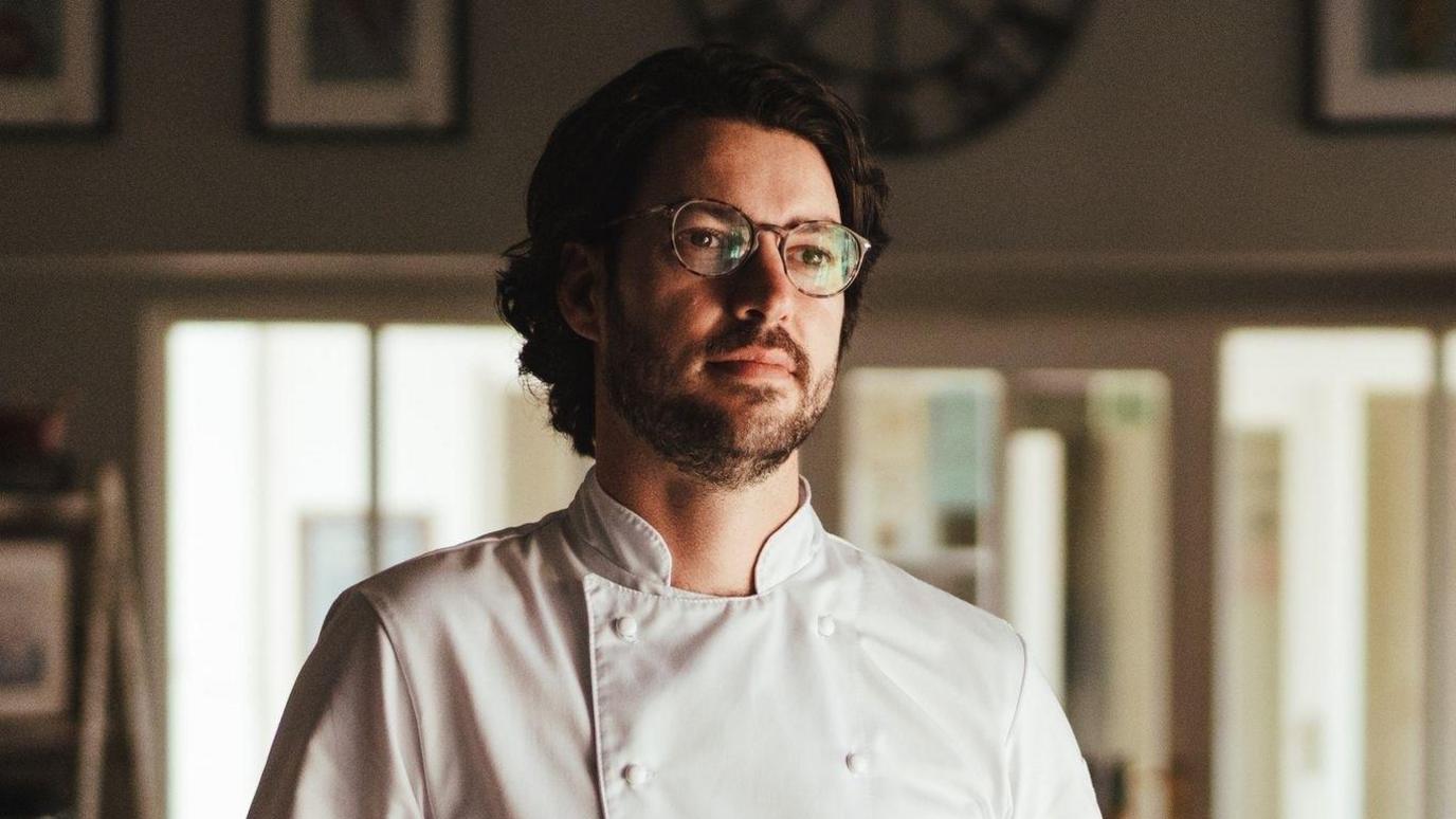 a man with glasses and curly black hair stands in a kitchen wearing his chef whites