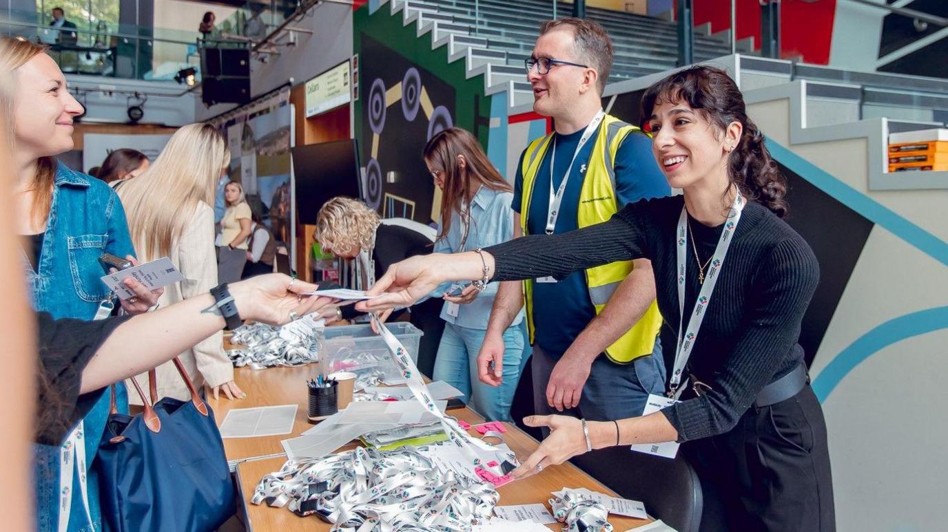 A man and a woman smile as they stand behind a table which is covered in lanyards as they welcome people to the Festival of Sustainable Business at Bristol Beacon